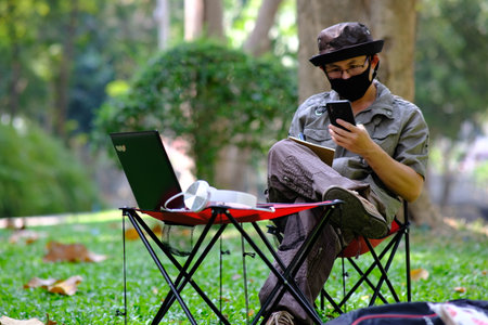 A man in a black protective mask is sitting on a chair and using a mobile phone in the park.