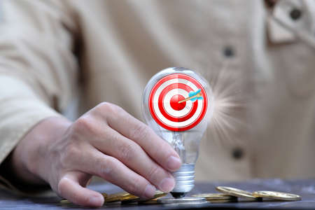 A person holding a lightbulb with a target on the background of coinsの写真素材