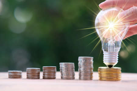 Businessman hand holding light bulb with money coins, Stacking coins on table with green bokeh...