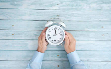 Man holding alarm clock on light blue wooden table, top view. Time management