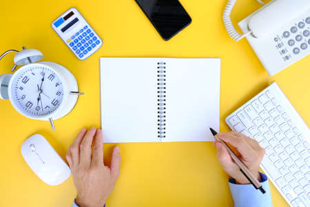 Top view of male hands writing in notepad and using computer keyboard on yellow background