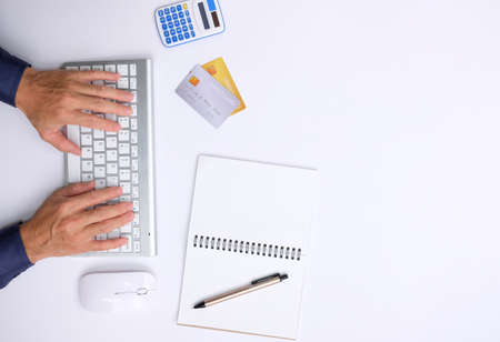 Top view of man using computer keyboard, credit card and notebook on white table. Online shopping...