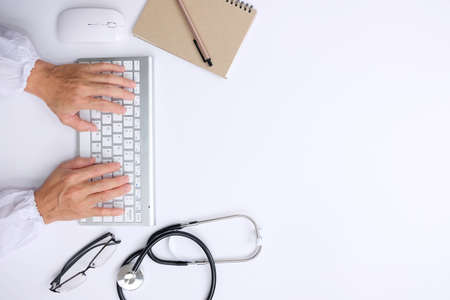 Top view of male doctor hands typing on computer keyboard with stethoscope and notebook on white...