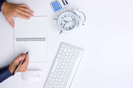 Top view of male hands writing in notebook on white table with alarm clock and calculatorの写真素材