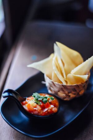 Tomato salsa and corn chips on wood table , Fresh tomato salsa with serrano pepper, red onion, green onion, lime, and cilantro in black dishの写真素材
