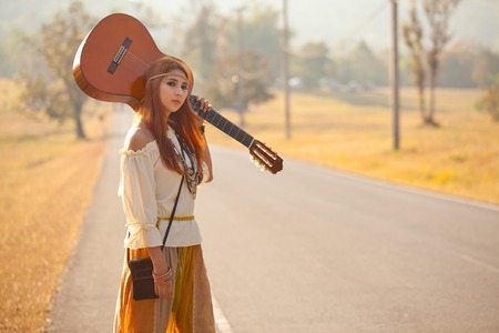 Hippie girl with guitar hitchhiking on countryside roadの写真素材