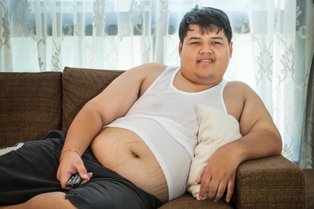 Overweight asian guy sitting on the couch with remote in hand trying to watch some TVの写真素材