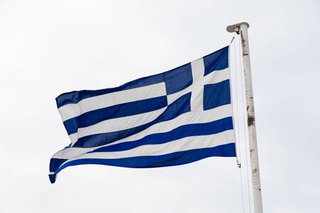 The Greek flag flying in the wind on a white flag pole.
