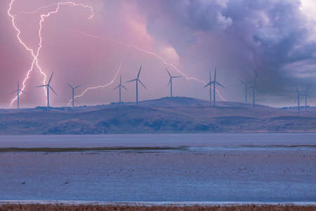 Photograph of large Wind Turbines in a row along a hill ridge line around a large lake during a lightning stormの写真素材