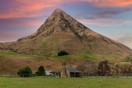 Photograph of an old small stone farm cottage in a lush green agricultural field in front of a large hill on the South Island of New Zealandの写真素材