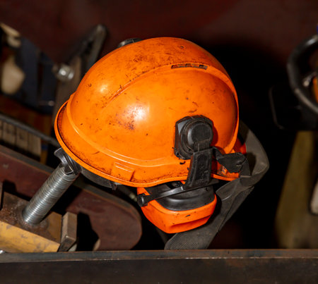 Photograph of an old and dirty orange safety hard hat with hearing protection laying on a metal...