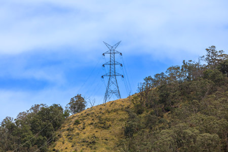Photograph of a large electricity Transmission Tower on a grassy hill against a bright blue sky in...