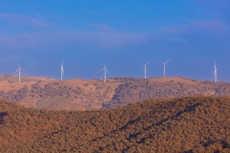 Photograph of a line of Wind Turbines standing on a large rural hill against a vibrant blue sky in...