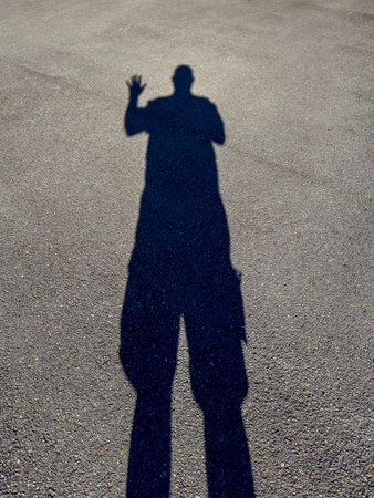 Photograph of a large black shadow on the ground of an asphalt carpark of a standing person due to a bright sunlight shining from behind them.の写真素材