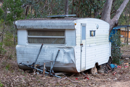 Photograph of an old rusty and destroyed caravan that has been parked and left on a side road adjacent to other discarded items in a large forest area.の写真素材