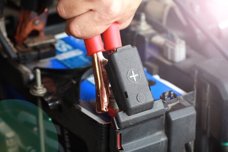 Close up of car battery anode cap with men's hand holding red electricity through cables for...