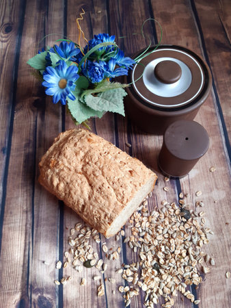 a wooden table top with brown stoneware dishes on it, half a loaf of bread next to spilled seeds of spelled, sunflower and pumpkin seeds. At the back, a tiny pot with blue margaritasの写真素材