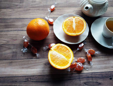 oranges are scattered and candies on a wooden table. A cup of tea and a teapot are standing