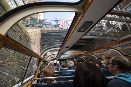 Amsterdam, Netherlands - April 20, 2017:A tourboat is taking tourists around the canals in Amsterdam. The boat is going under a bridge. Classic architecture and boats flank the canal.のeditorial素材