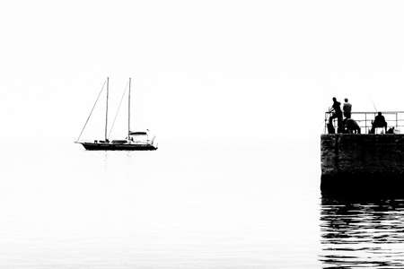 Silhouettes of fishermen on the pier on the background of the silhouette of a yacht on the calm sea. Black and white photo. High quality photoの写真素材