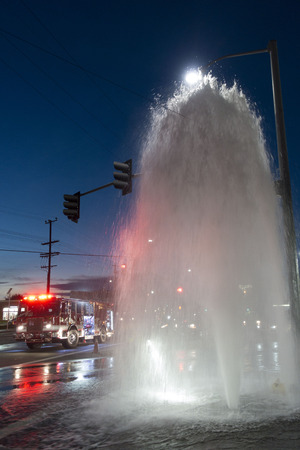 A fire truck arrives at an intersection filled with water from a broken fire hydrant shooting water twenty-five feet into the air beneath a street lamp at sunset.のeditorial素材