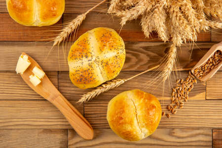 Three buns lying on a wooden table next to the grains and ears of wheat. Next to it is a wooden knife with butter. View from above.の写真素材