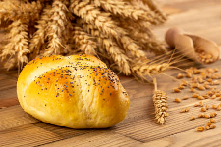 A close-up of a bun lying on a wooden table next to grain and ears of wheat.の写真素材
