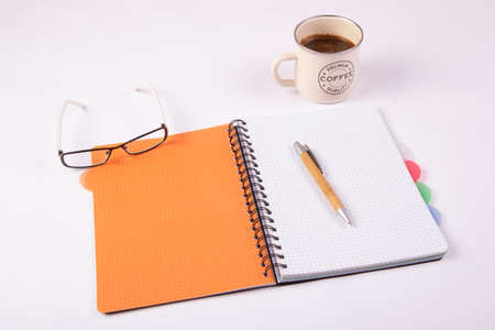 Opened orange notebook with a pen, glasses and a mug of coffee on a white backgroundの写真素材
