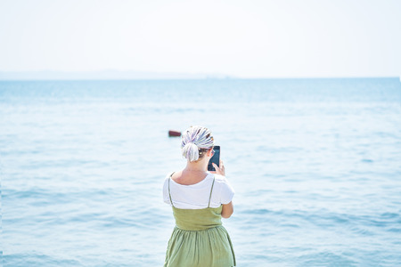 Young woman with multi-colored dreadlocks in a light cotton sundress stands on the beach at the water's edge and take pictures of the sea at her smartphone. View from the backの写真素材