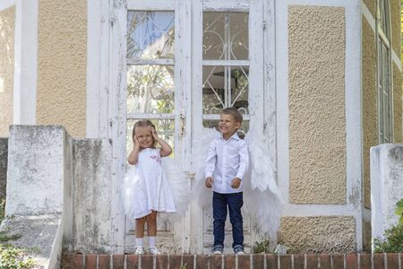 Two blond little children, brother and sister, dressed as angels with white wings, stand in front of a closed glass door to a stone house, stone steps are visible belowの写真素材