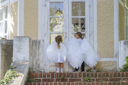 Two blond little children, brother and sister, dressed as angels with white wings, stand in front of a closed glass door to a stone house, stone steps are visible belowの写真素材