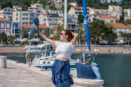 Young woman tourist in a hat, sunglasses, blue skirt and white blouse walking on the promenade of a seaside town. Residential buildings and a yacht moored at the pier are visible behind herの写真素材
