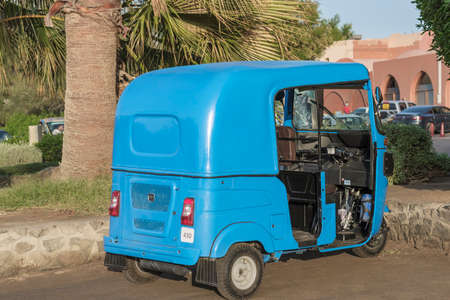 El Gouna, Egypt, November 08, 2021: Blue Tuk-tuk without a driver close-up. It is standing on the street by the road under a palm tree. In the background there is a parking lot for carsのeditorial素材