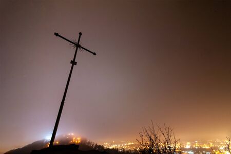 The Christian cross rises high mountain against the backdrop of a foggy night city.の写真素材