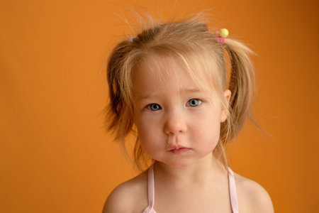 A little girl at the age of one and a half years sits on her lap and cries on a yellow background....