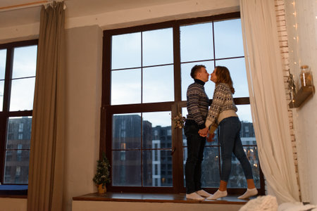 A loving couple in knitted sweaters stand by the hand against the background of a large window on the windowsill.の写真素材