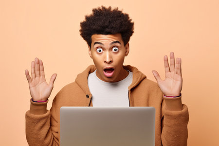 Surprised African teen guy holding laptop pointing at computer advertising wow offer. Amazed ethnic teenager student boy using pc feeling shocked isolated on beige backgroundの素材