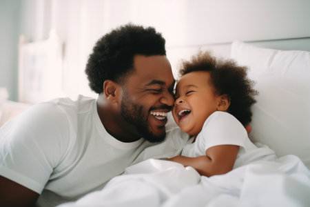 Happy loving young Black dad holding adorable mixed race baby daughter having fun in bed at home. Smiling African father playing with cute funny infant child girl waking up in bedroom in the morningの素材