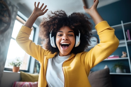 Happy funny gen z hipster African American teen girl wearing headphones dancing at home, listening music on mobile phone, having fun feeling funky moving in living roomの素材