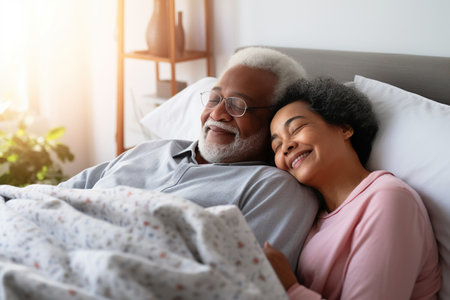 Happy senior biracial couple lying in bed and embracing at homeの素材
