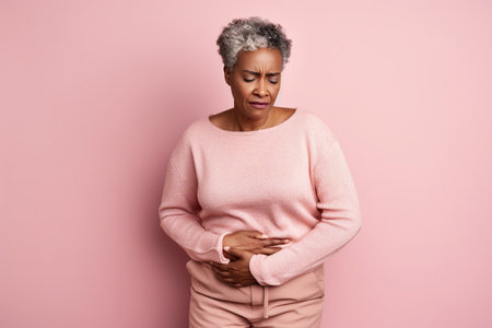 Old African American woman holding stomach with discomfort from gastritis on pink backgroundの素材
