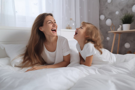 Cheerful caring mom lying at kid on double bed before yawning toddler girl falling asleep, talking with child, laughing, relaxing on spacious comfortable mattress with white sheetの素材