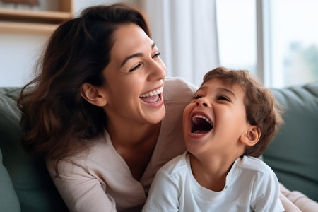 Happy young Latin mother and little son having fun on cozy couch in living room, overjoyed mum and preschool child tickling, cuddling, enjoying leisure time together,の素材
