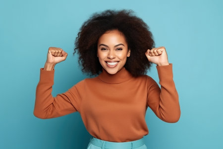 Pretty young African American girl raise fist celebrate hold gadget dressed stylish brown outfit isolated on blue color backgroundの素材