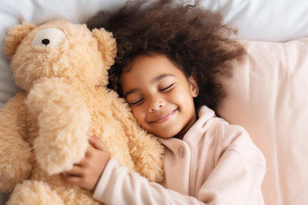 Top view of cute little preschooler African American girl lying cuddling with favorite fluffy toy waking up in cozy home bedの素材