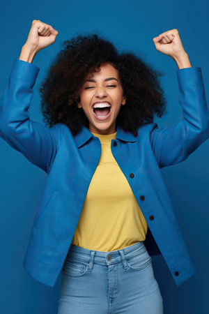 Photo of overjoyed cheerful African American woman dressed stylish clothes celebrate success raise hands fists isolated on blue color backgroundの素材