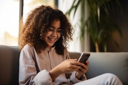 Happy young Latin woman sitting on sofa holding mobile phone using cellphone technology doing ecommerce shopping, buying online, texting messages.の素材