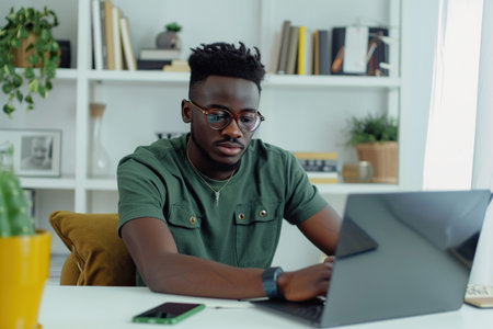 Young African American man wearing jeans and green t-shirt sitting working in laptop in stylish white home officeの素材