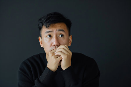 Nervous Asian man and biting nails in studio with oops reaction to gossip on black background. Mistake, sorry and male overwhelmed by fake news, drama or secret with regret, shame or awkwardの素材