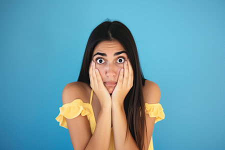 Nervous Latin woman in studio with oops reaction to gossip on blue background. Mistake, sorry and female overwhelmed by fake newsの素材